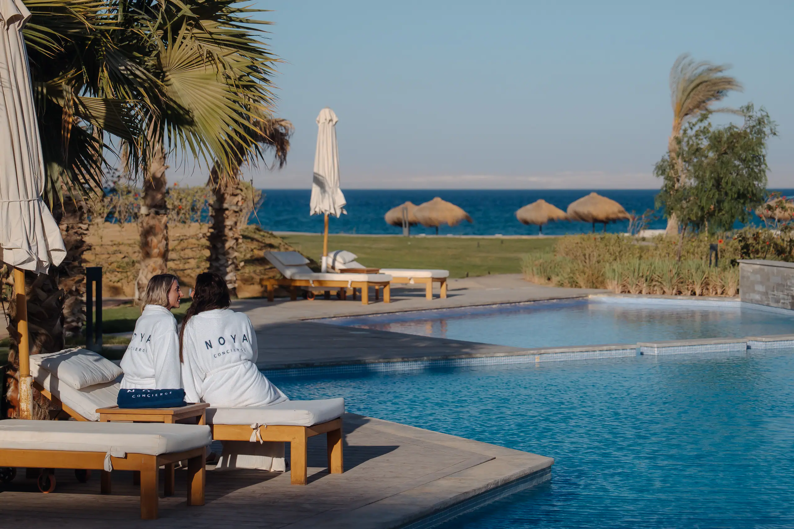 Two guests in NOYA robes by a pool in El Gouna, beach parasols and the Red Sea beyond