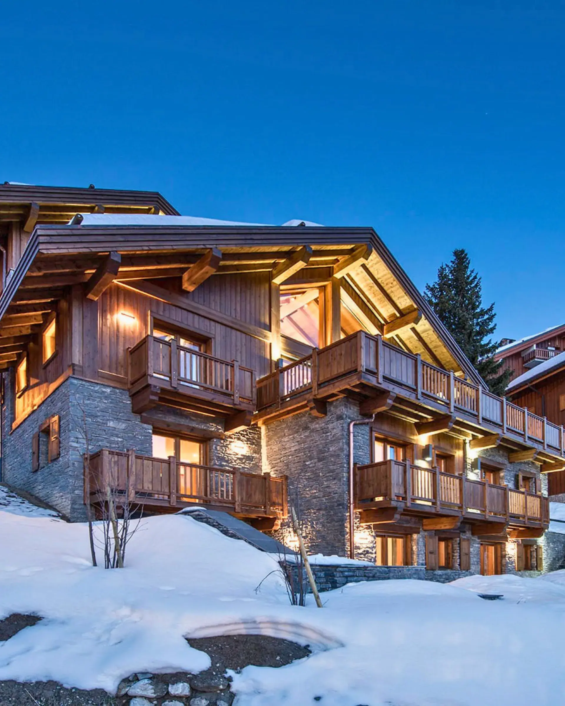 A timber-and-stone chalet at blue hour, its balconies lit, deep snow around it
