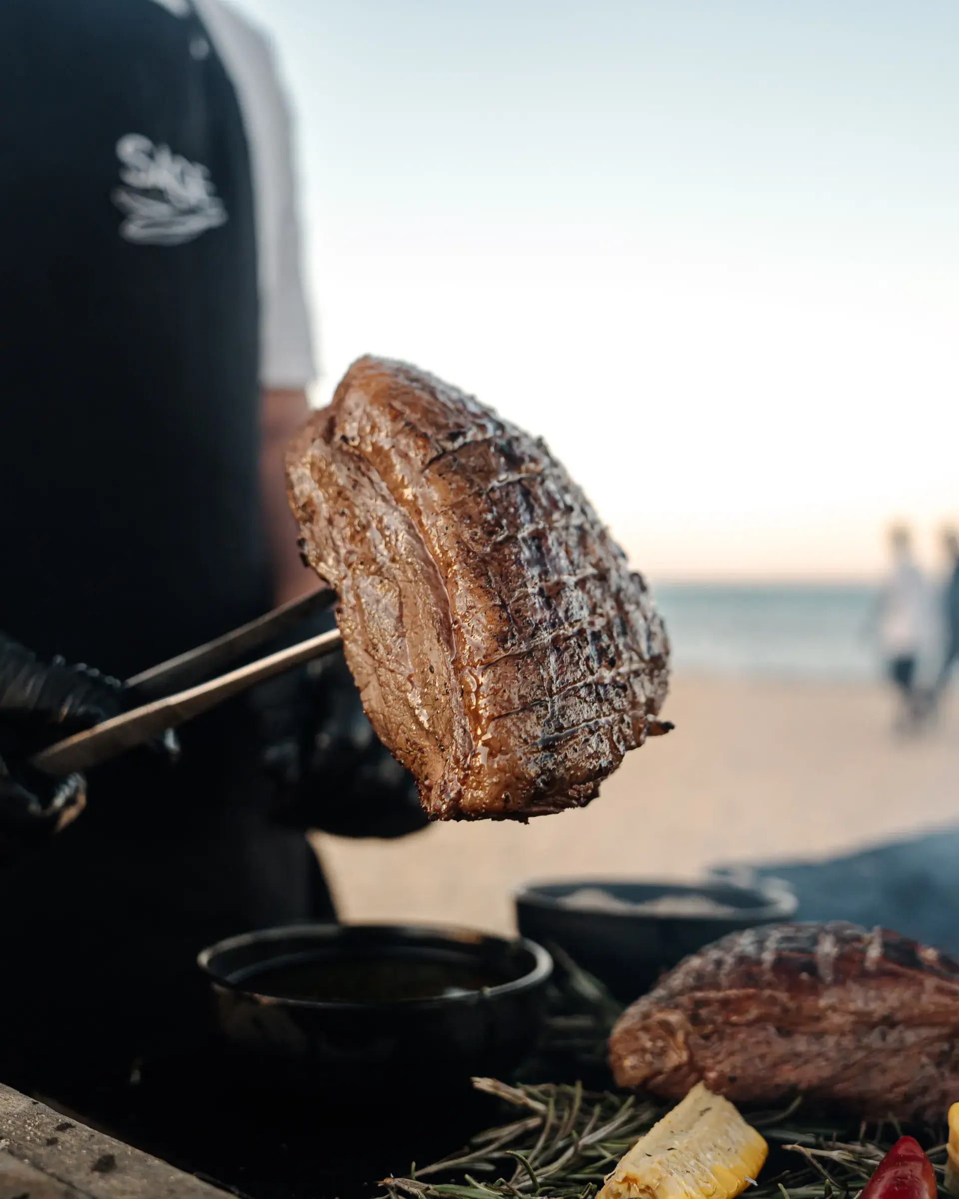 A chef lifting grilled picanha from the fire on the beach at golden hour, the sea behind