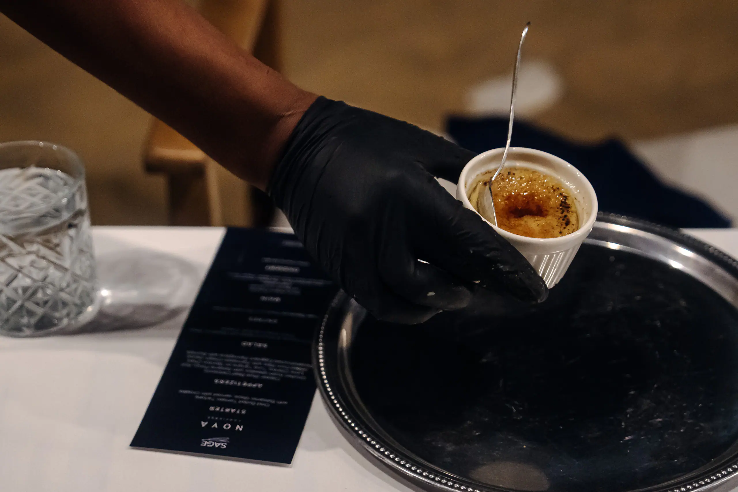 A gloved hand serving crème brûlée beside a NOYA × Sage menu